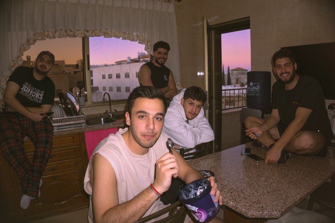 The same group of friends in a kitchen at sunset, one of them in a white shirt eating from a tub of ice cream with a spoon.