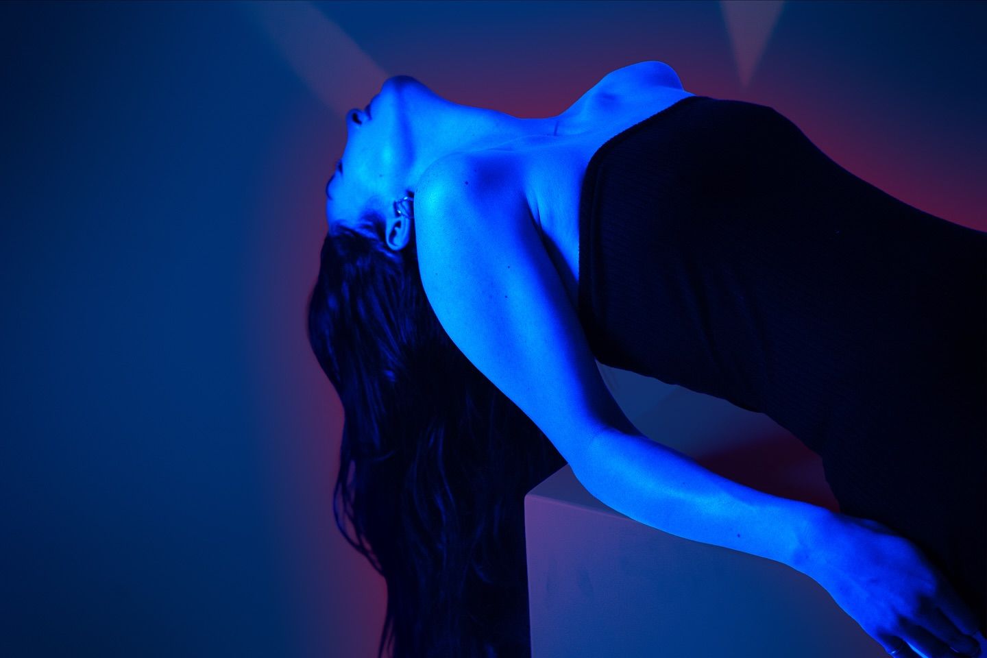 A woman arches backward over a white block, her bare shoulders and face lit in cool electric blue, dressed in black.
