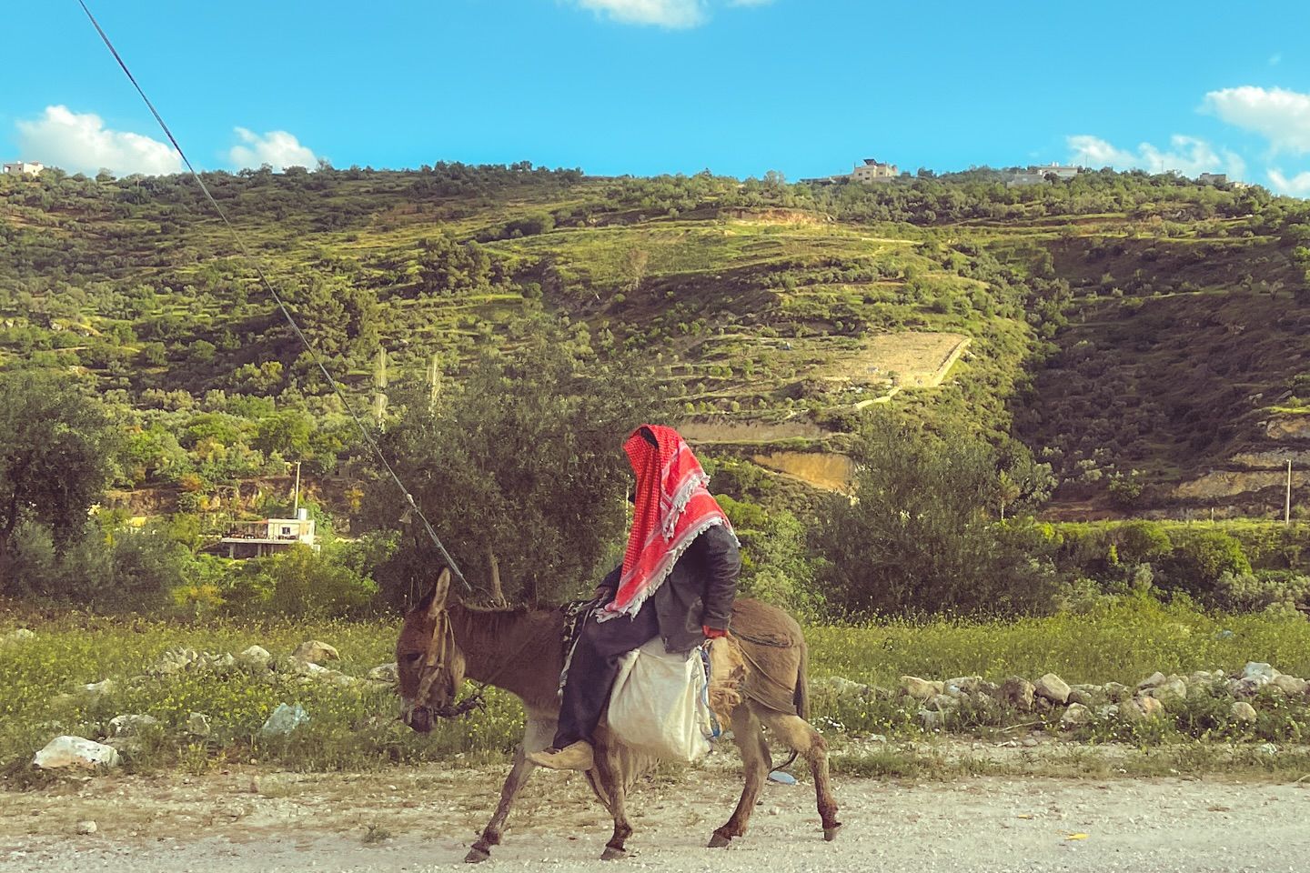 A person in a red-and-white checkered headscarf rides a donkey along a dirt path, terraced green hills and scattered houses behind them.