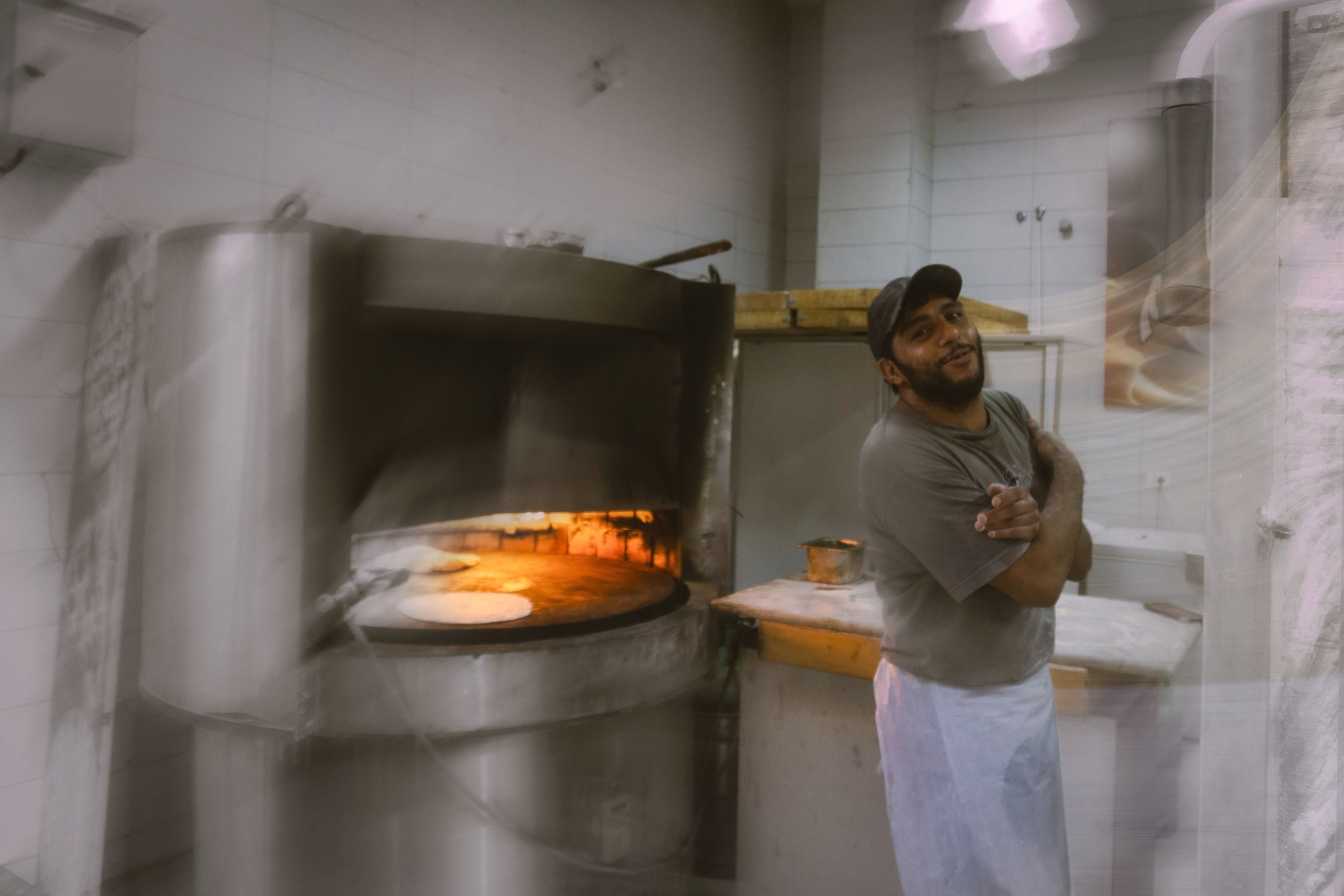 A man in a cap stands smiling beside a wood-fired oven with flatbread baking on its round stone, a double-exposure effect over the scene.
