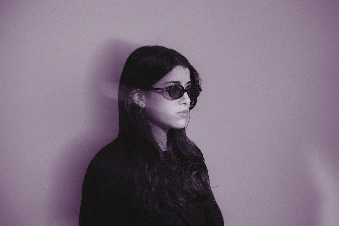 A profile portrait of a woman in dark sunglasses from a slightly different angle, same violet-toned backdrop.