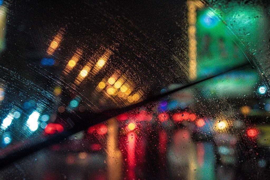 A blurred night cityscape seen through a rain-streaked window, car lights and a green-lit sign smeared into trails of colour.